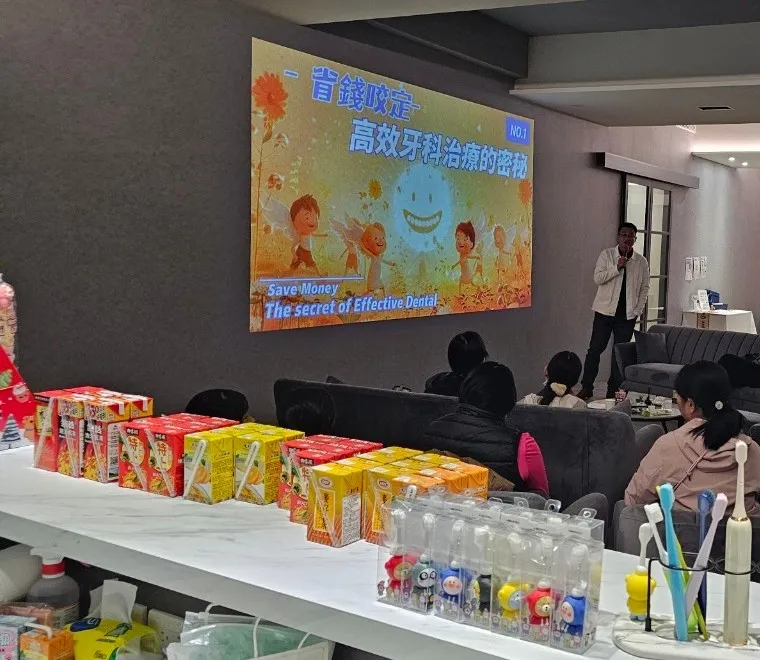 講座現場,講者手持麥克風站在投影幕旁,簡報主題是省錢又高效的牙科治療;前方檯面擺著成排的鋁箔包飲料與牙刷,聽眾背對鏡頭坐在沙發區