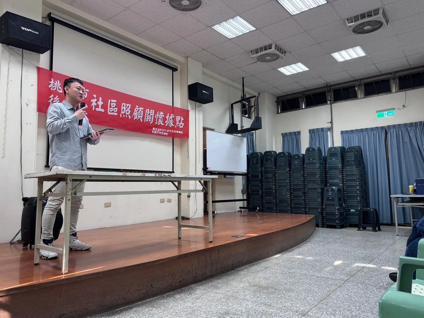 講師站在據點的舞台前,身後是「桃園市中壢區後寮里社區照顧關懷據點」的布條