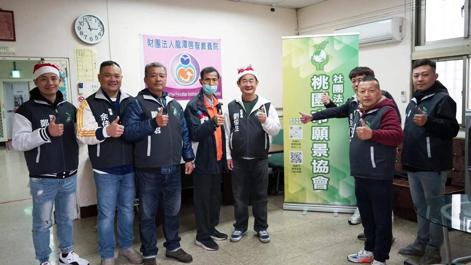協會理監事戴著聖誕帽，在龍潭啟智教養院室內與協會立牌合影