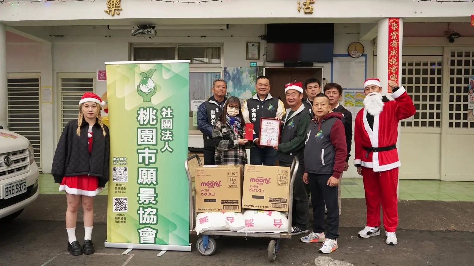 協會理監事戴著聖誕帽，在樂活育幼院門口與推車上的捐贈物資和協會立牌合影
